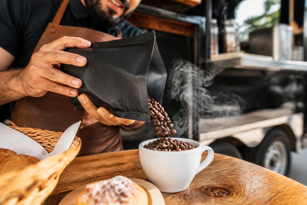 Close-up of freshly roasted coffee beans in various roast colors being poured into a white ceramic cup with steam rising, showcasing whole bean quality and roast color variation