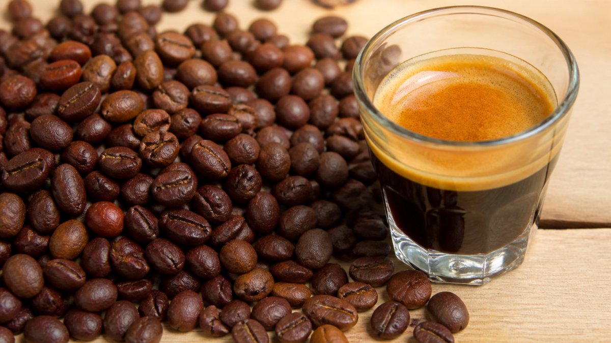 Roasted coffee beans in deep brown and red tones scattered next to a glass of fresh espresso on a wooden surface.
