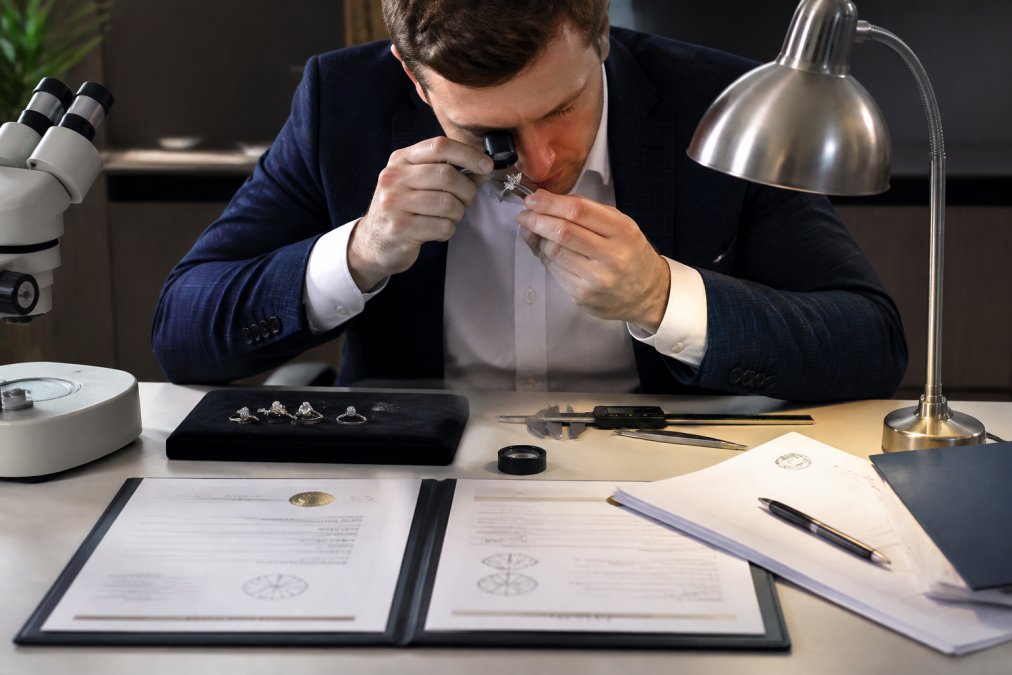 Professional appraiser examining a diamond ring with a jeweler's loupe and precision measurement tools on a workspace, with certification documents, appraisal forms, and lighting equipment visible