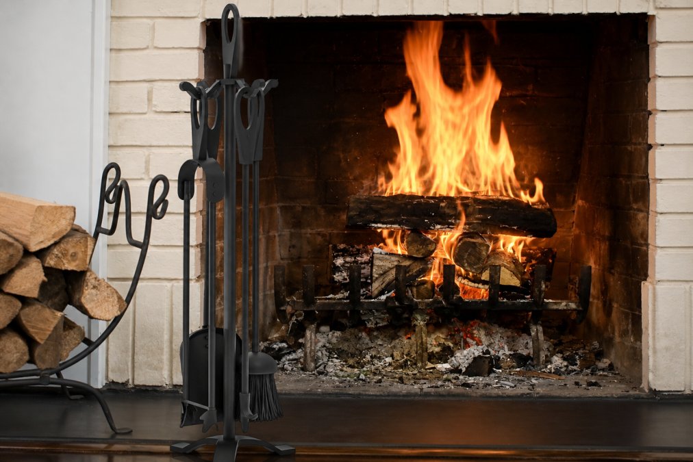 Close-up of premium wrought iron and black iron fireplace tool sets arranged on a traditional hearth, showing rich metal textures, patina development, and contrasting finishes under warm firelight