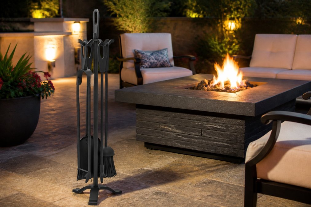 Elegant outdoor patio setting with premium fire table as centerpiece, coordinated black iron accessories arranged nearby, comfortable seating arranged for entertaining, warm evening lighting casting shadows across stone patio
