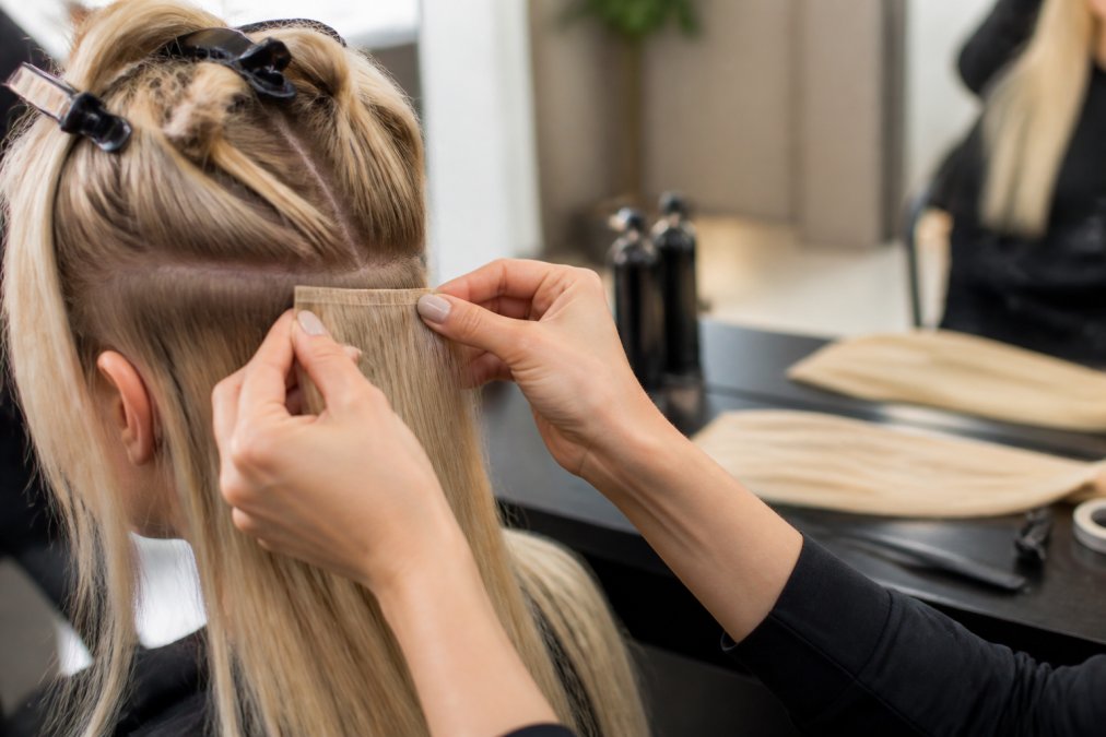 Close-up of a hairstylist's hands sectioning fine blonde hair and positioning a flat weft extension close to the scalp, natural salon lighting, focus on the seamless root blend