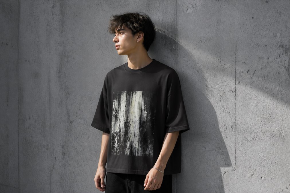 A person wearing an oversized heavyweight cotton graphic tee standing against a plain concrete wall, fabric falling straight from the shoulder without clinging, soft daylight from the side