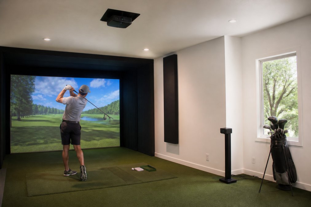 A golfer mid-swing inside a spacious home simulator room, overhead launch monitor mounted on the ceiling, large impact screen glowing with a virtual fairway ahead