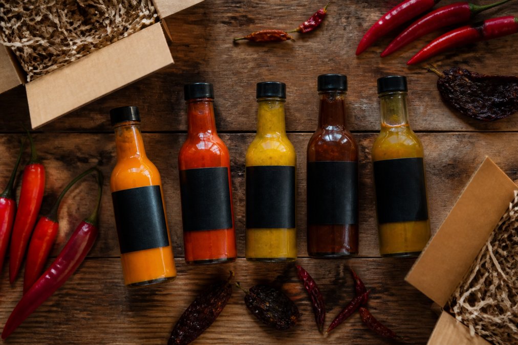 Flat-lay of five artisanal hot sauce bottles with distinctive labels arranged on a worn oak surface, surrounded by fresh red chilli peppers, dried chipotle pods, and open kraft-paper gift boxes under warm directional lighting