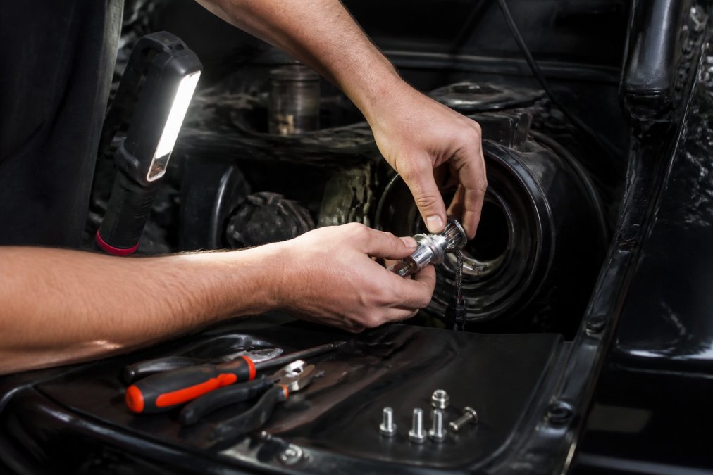 Professional technician installing LED bulb into classic car's headlight housing, carefully positioning the bulb and rotating the base for beam alignment, hands visible with work light illuminating the engine bay