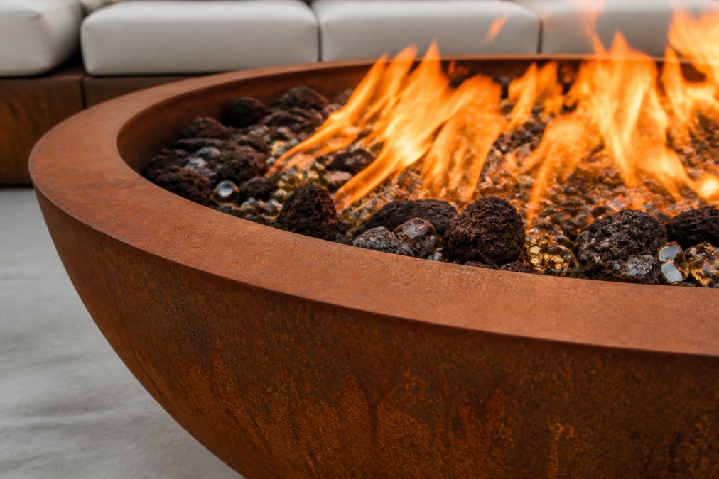 Close-up detail of a handcrafted Corten steel fire pit showing the natural rust patina finish and industrial craftsmanship, with fire glass and lava rock visible through the flames