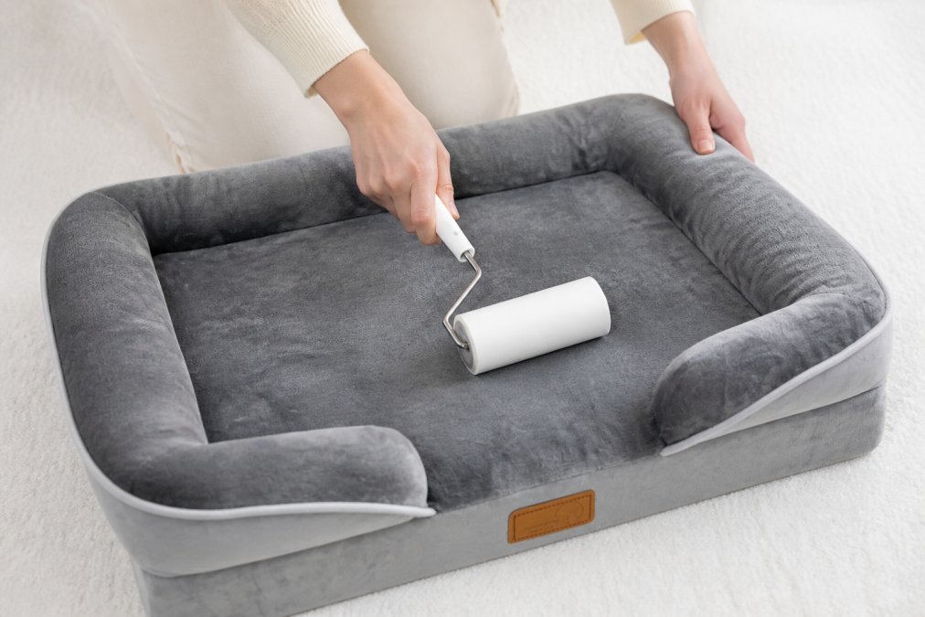 Close-up of hands cleaning a pet bed cover with a lint roller, showing the bed in clean, unused condition on a light background
