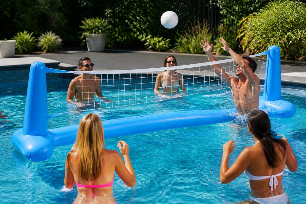 Adults in a swimming pool actively playing with inflatable volleyball equipment, some passing a ball while others watch, with bright sunny conditions and water splashing