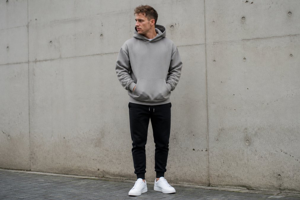 Person wearing a premium oversized heavyweight hoodie in neutral grey, styled with fitted black joggers and white minimalist sneakers, standing in natural daylight against a neutral urban wall, showing how the structured fabric drapes cleanly from the shoulders