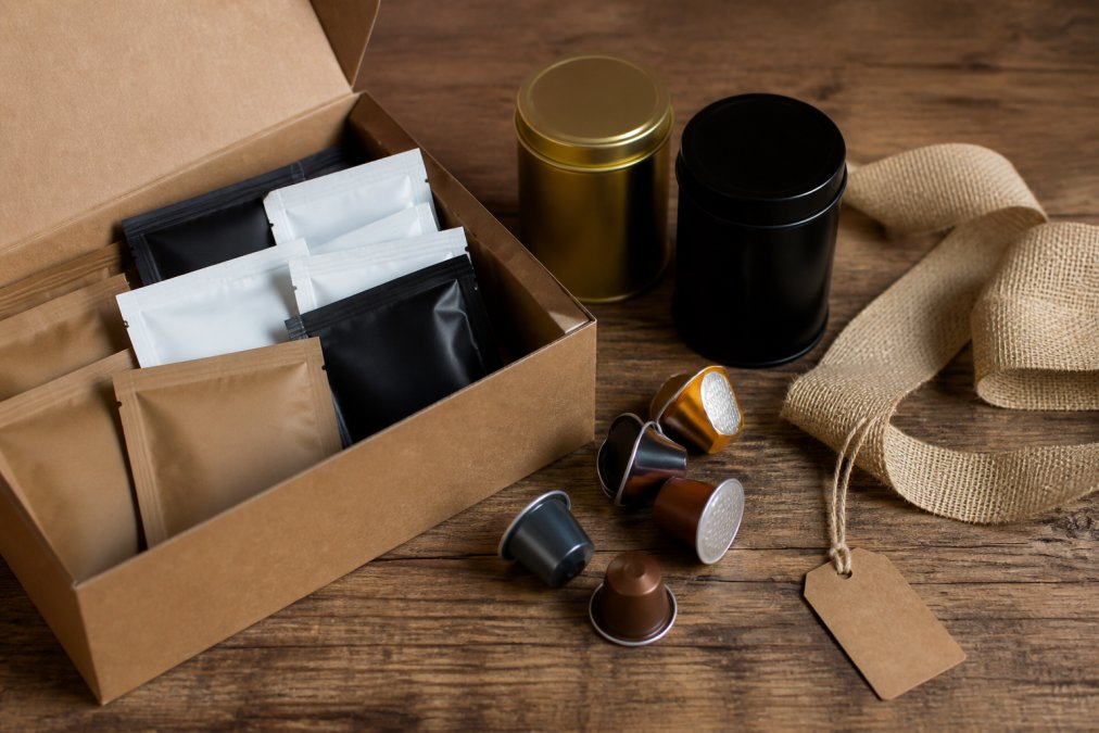 A flat lay of Cenia's Coffee sample packs, tea tins, and coffee capsules arranged on a rustic wooden table, with a burlap ribbon and a small kraft gift tag beside them, soft natural window light