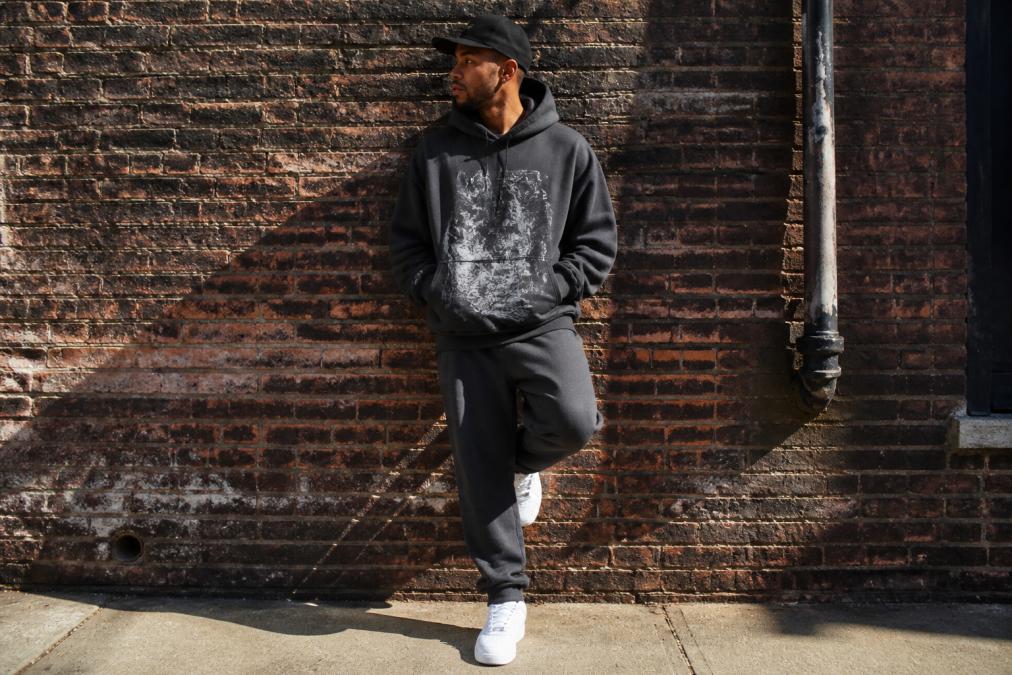 A stylish man in his late 20s wearing a premium oversized graphic hoodie, matching track pants, and clean white sneakers, posed against a weathered brick wall in soft natural light with deep shadows