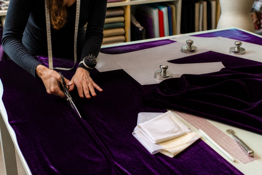 A seamstress at a well-lit sewing table carefully cutting deep purple velvet fabric with sharp shears, the fabric grain clearly visible, with interfacing, pattern weights, and sewing tools arranged neatly nearby