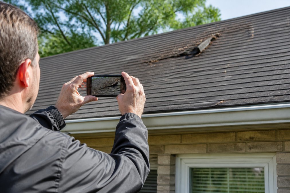 Homeowner photographing roof damage from ground level with smartphone, capturing close-up view of damaged shingles, granule loss, and storm debris scattered on roof surface in natural daylight