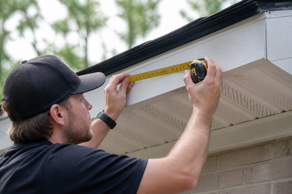 Close-up of homeowner examining roofline fascia board and soffit with measuring tape, showing mounting points and surface preparation areas with natural daylight