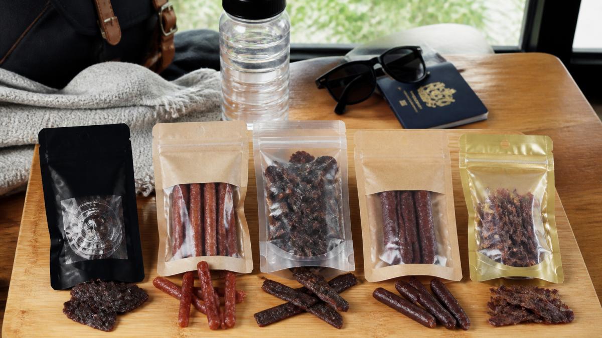 Flat-lay arrangement of five different preservative-free meat snack products displayed on a wooden cutting board with travel accessories including a passport, sunglasses, and water bottle in the background, natural daylight from window