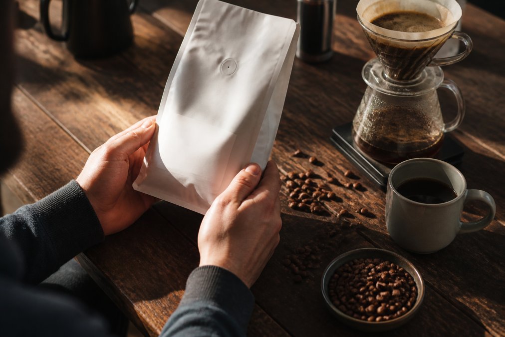 Hands holding a specialty coffee bag displaying roast date and single-origin information, with a pour-over dripper, whole beans scattered on dark wood, and a freshly brewed cup of coffee in warm afternoon light