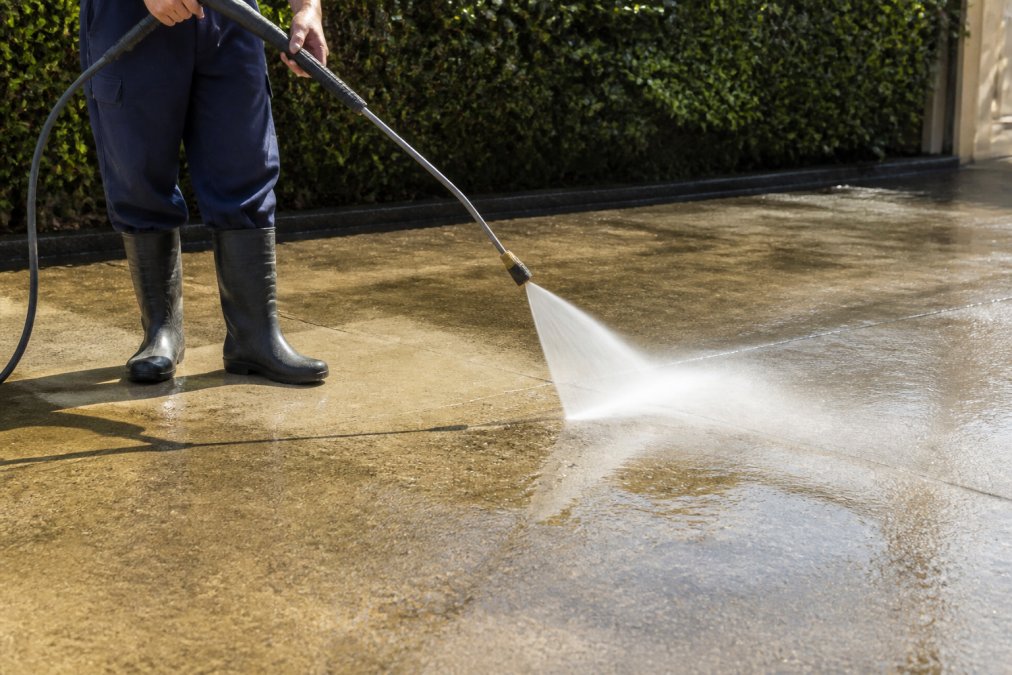 Close-up of a person in work boots and long trousers correctly angling a pressure washer wand at approximately 45 degrees from a concrete driveway surface, with a visible wide fan spray pattern and water running across the concrete in bright afternoon sunlight
