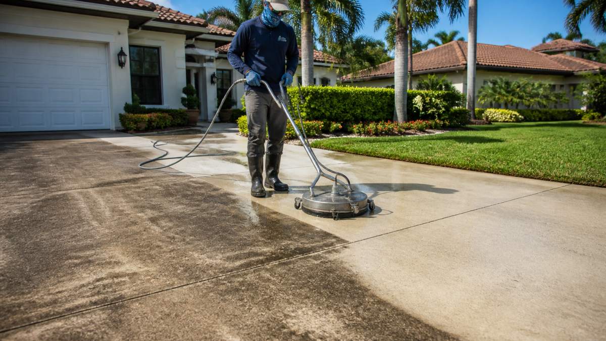 A technician in protective gear using a surface cleaner attachment on a concrete driveway, with a visible clean stripe beside a dirty section, bright afternoon sunlight