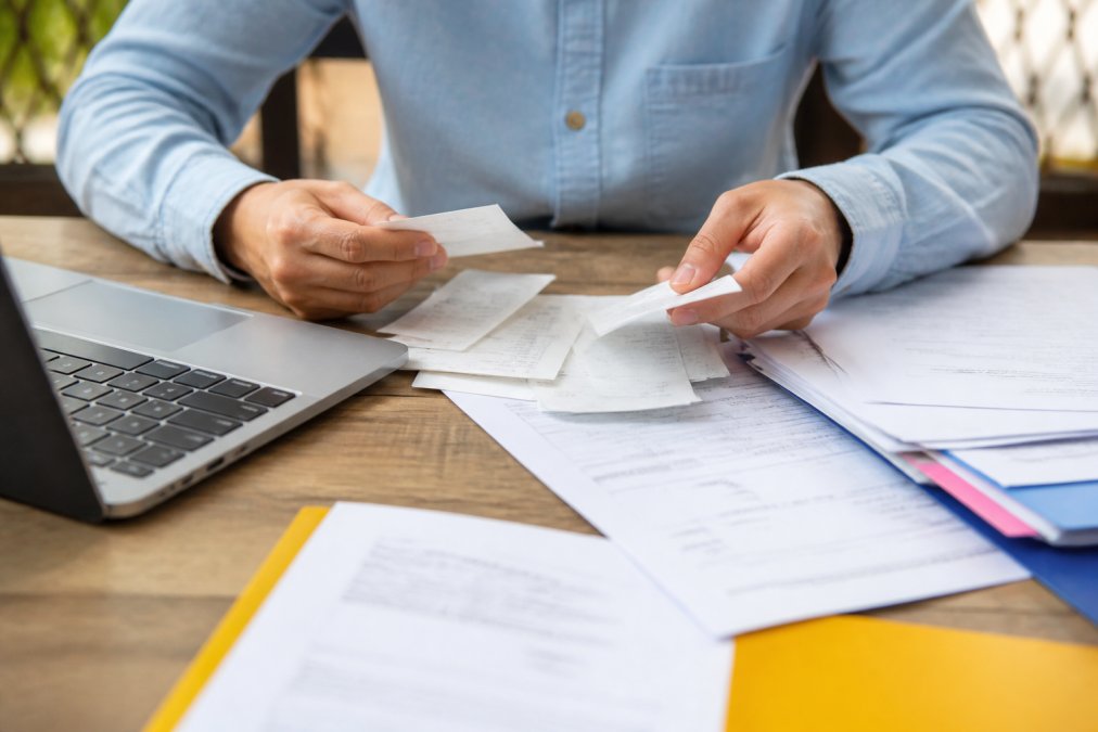 Plan rapproché des mains d'une personne organisant des documents financiers, relevés bancaires et justificatifs sur un bureau avec un ordinateur portable