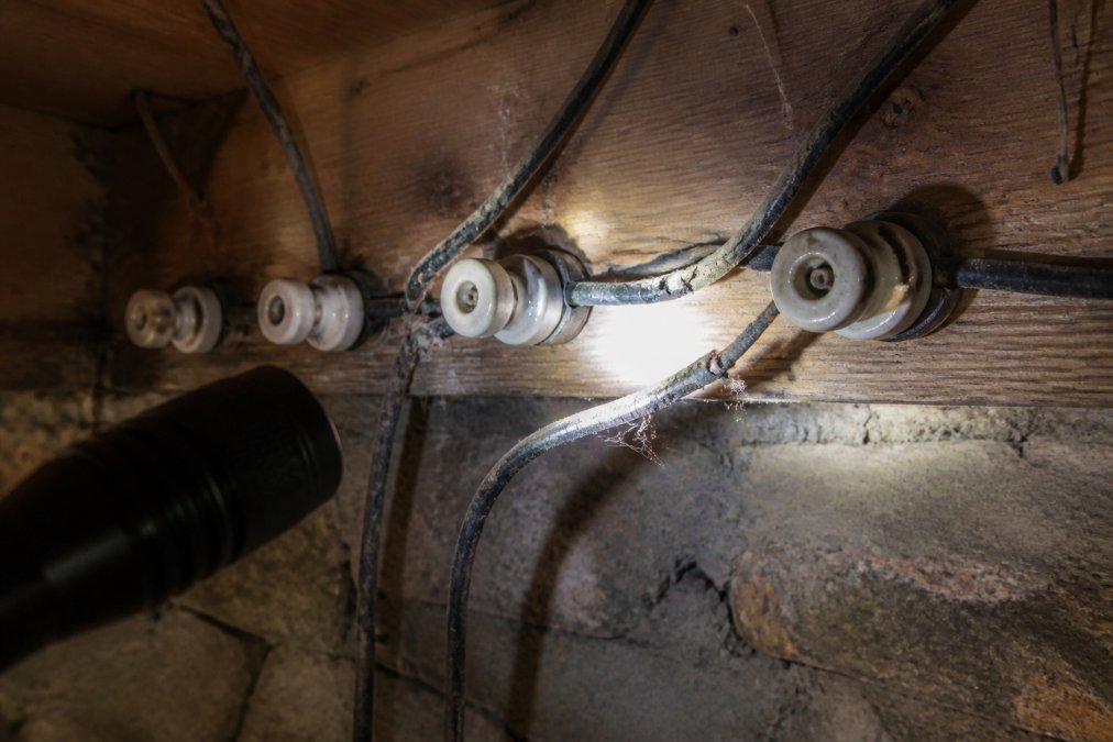 Close-up of an old knob-and-tube wiring system in a dimly lit basement, showing cracked insulation and ceramic knobs, with a flashlight beam highlighting the deterioration