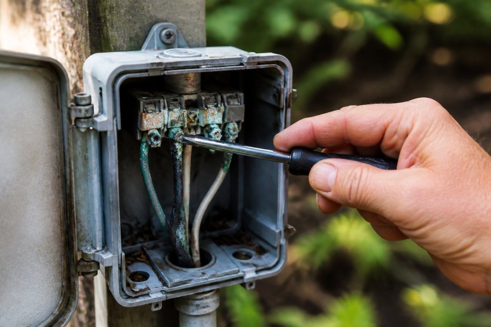 Close-up of an outdoor electrical junction box with visible green corrosion on copper wire connections and a technician's hand pointing to the corroded area with a tool