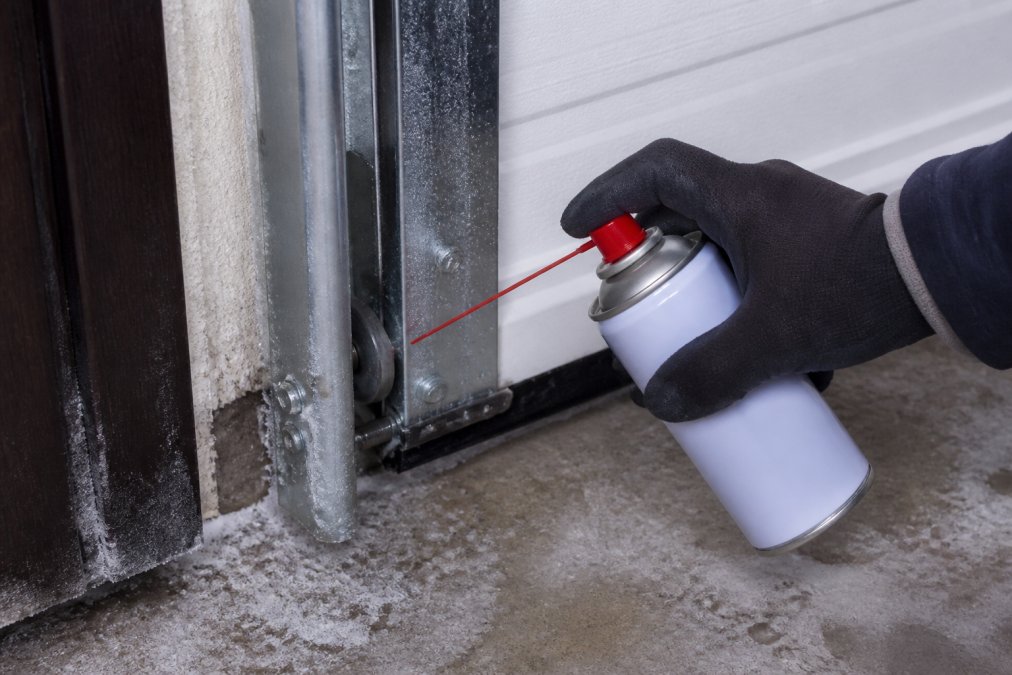 Gloved hand applying silicone lubricant to frosty garage door tracks to prevent garage door freezing during winter.