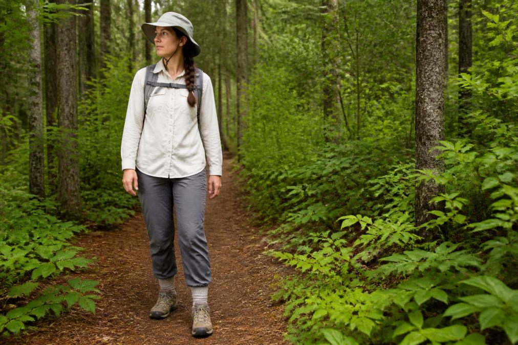 Hiker wearing light-colored long-sleeved shirt, long pants tucked into thick socks, and a wide-brimmed hat while standing on a forest trail surrounded by vegetation