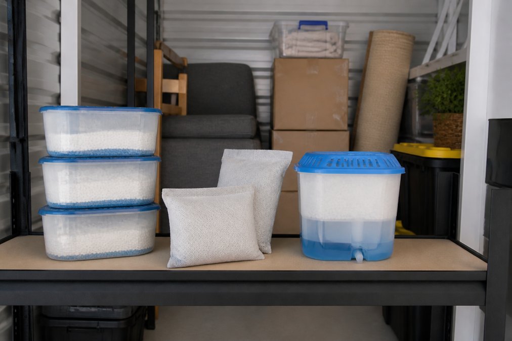 Close-up of various moisture absorber products including silica gel containers, desiccant bags, and calcium chloride dehumidifiers arranged on a shelf inside a clean, well-lit storage unit with stored boxes and furniture visible in background