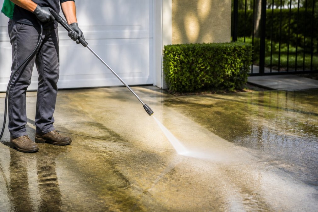 Professional demonstrating pressure washing technique on concrete driveway surface with high-pressure water stream removing visible mold and algae, showing water and debris being cleared from the surface in bright daylight