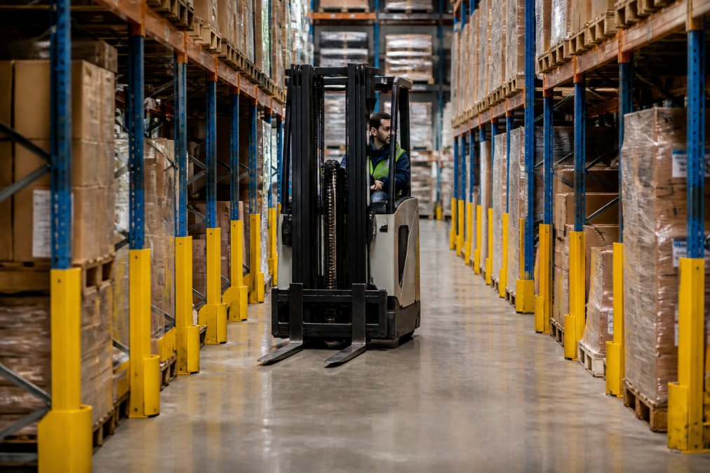 Warehouse operator carefully navigating a forklift through narrow aisles between pallet racking, with protective guards visible on column uprights in a busy retail storage facility