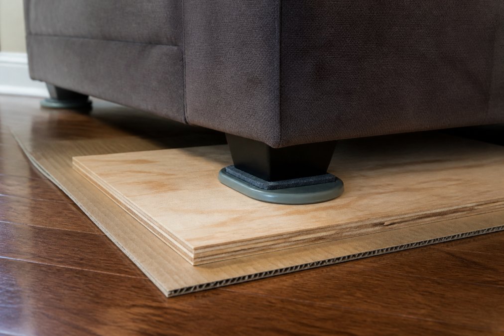 Close-up of furniture sliders positioned under a heavy sofa leg on hardwood flooring, with cardboard and plywood sheets layered underneath for multi-level floor protection in natural daylight