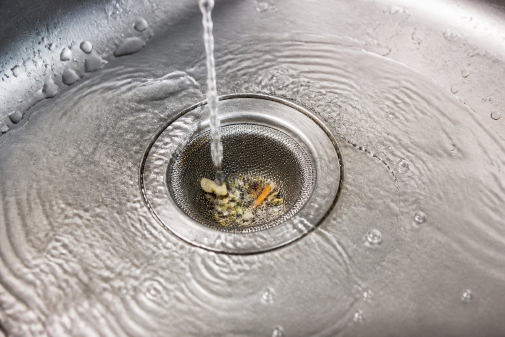 Close-up of a kitchen sink drain with a stainless steel strainer basket catching food particles, water running, bright natural light