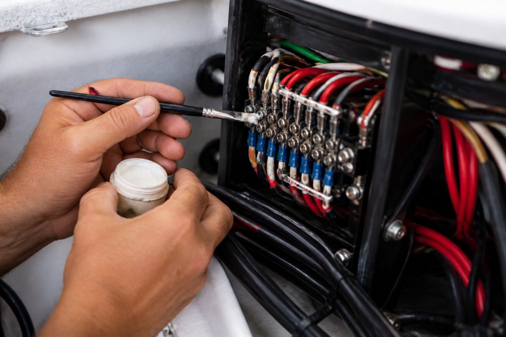 Technician's hands applying dielectric grease to marine connector pins on a boat's electrical panel with a small brush, showing protective coating being worked into terminal block gaps