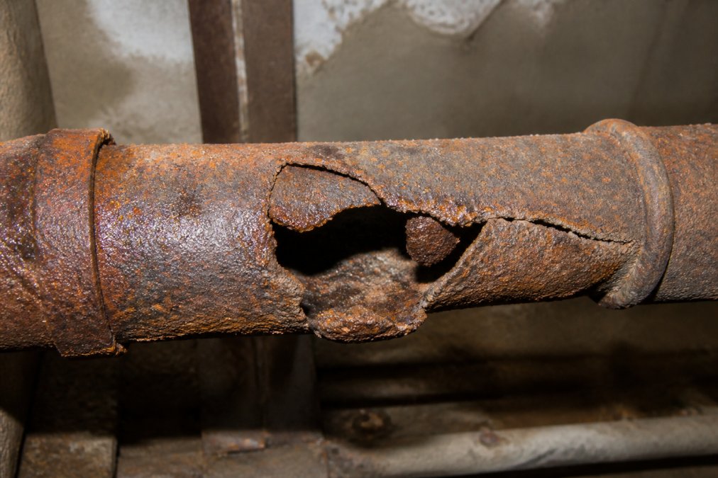 Close-up of severely corroded and fractured cast iron sewer pipe showing rust, deterioration, and structural damage in a basement setting with dim lighting