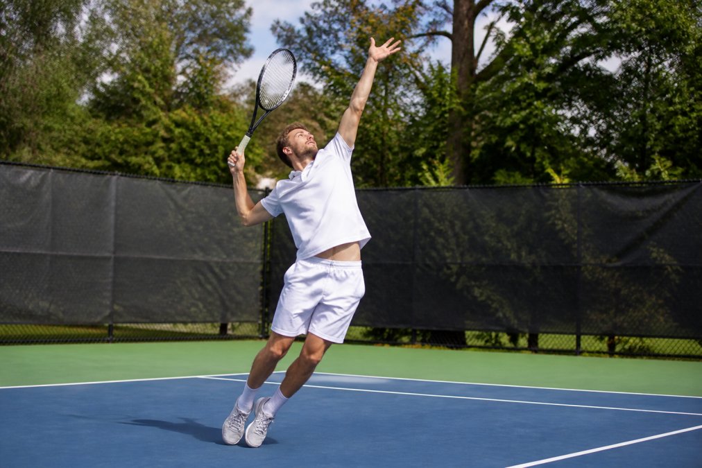 Tennis player mid-serve showing proper kinetic chain alignment with weight transferring from back leg through torso to shoulder, demonstrating correct posture and arm position with natural outdoor court lighting