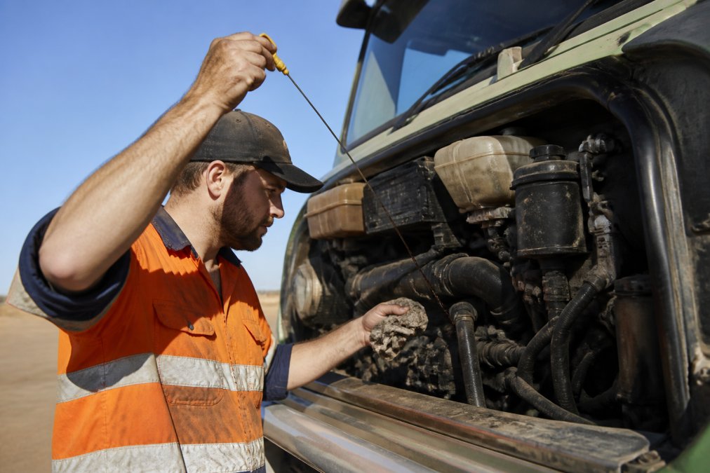 Mechanic checking engine oil level with dipstick on an agricultural truck in bright daylight, engine bay open with hands visible performing the inspection