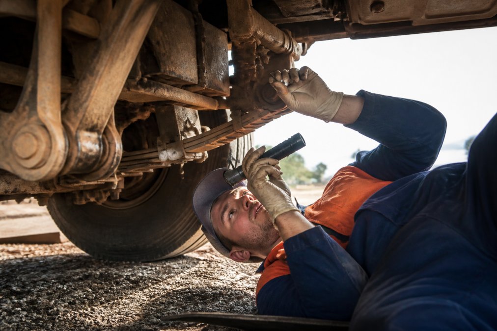 Farm truck undercarriage being inspected by mechanic with torch examining suspension joints and lubrication points in daylight