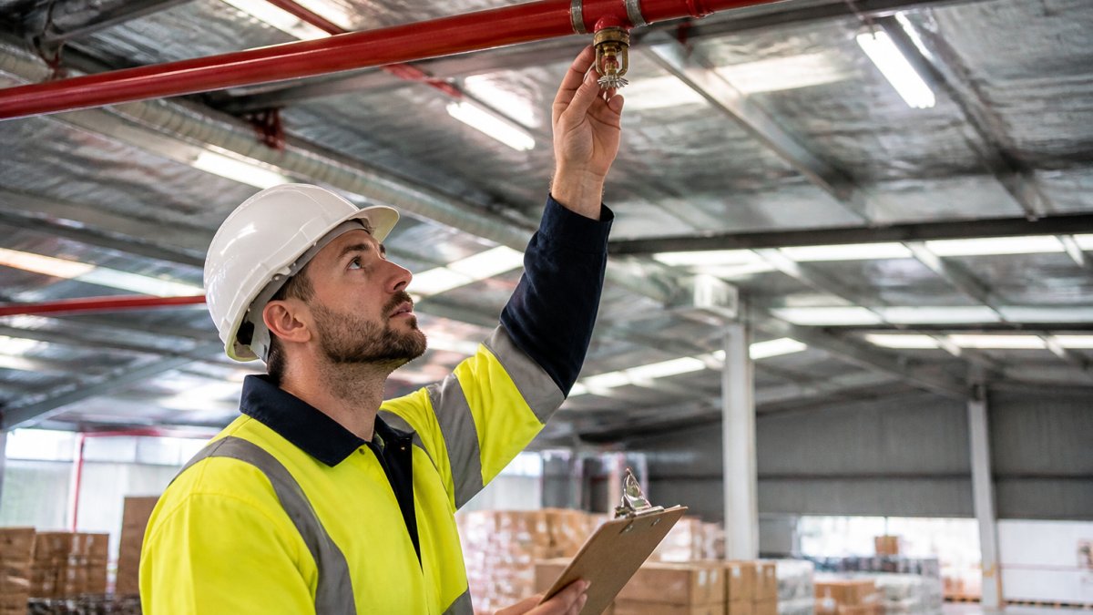 A fire protection technician in high-visibility gear and a hard hat inspecting a red commercial sprinkler head and piping in a warehouse ceiling, holding a clipboard under bright industrial lighting