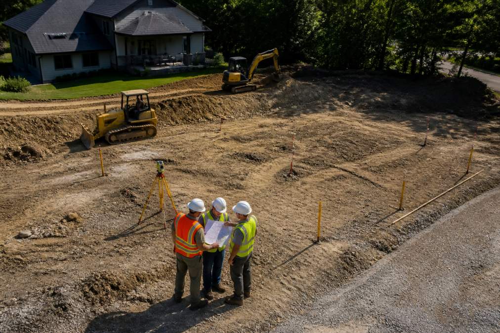 Overhead view of a residential construction site with survey markers, grading equipment, and crew members reviewing site plans on clipboard