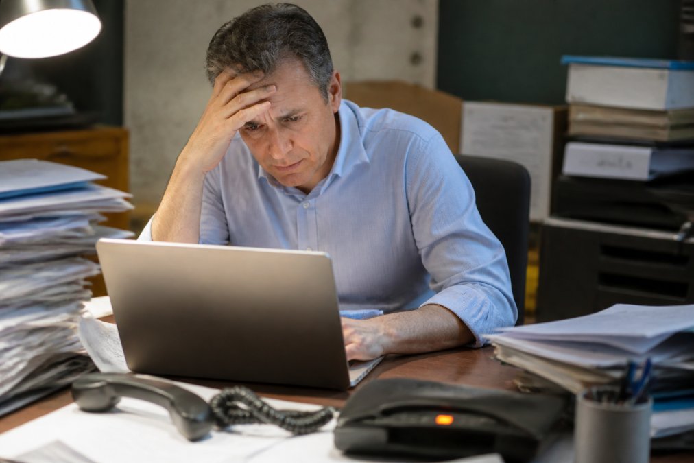 A small business owner staring at a laptop screen with a worried expression in a modest office, stacks of paperwork and a ringing phone nearby, harsh overhead lighting