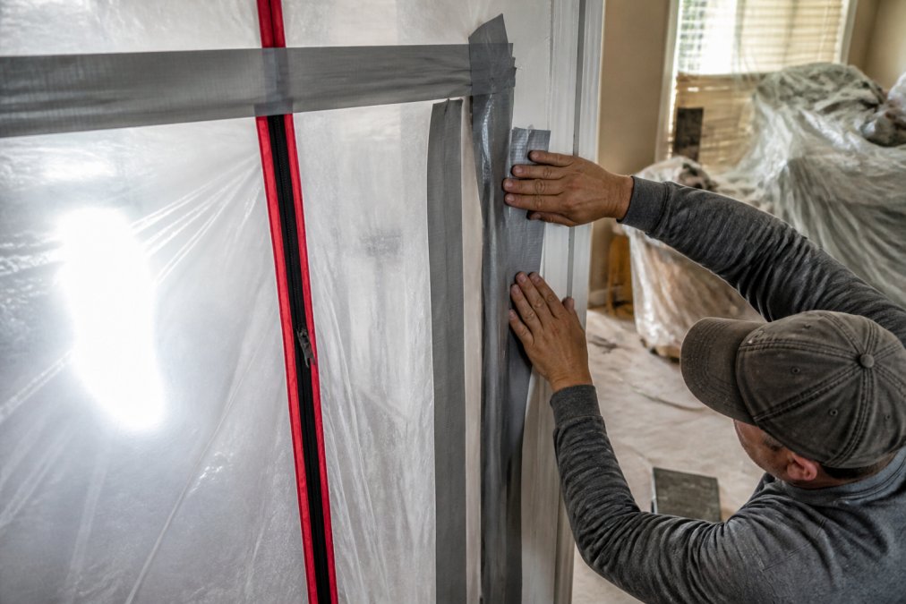 Close-up of plastic sheeting sealed with duct tape along doorway frame, with zip wall zipper visible and dust containment barrier fully installed during active renovation