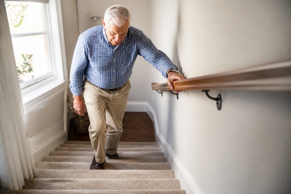 A senior man gripping a sturdy wooden handrail while carefully descending a carpeted indoor staircase, with bright natural light from a window illuminating the steps