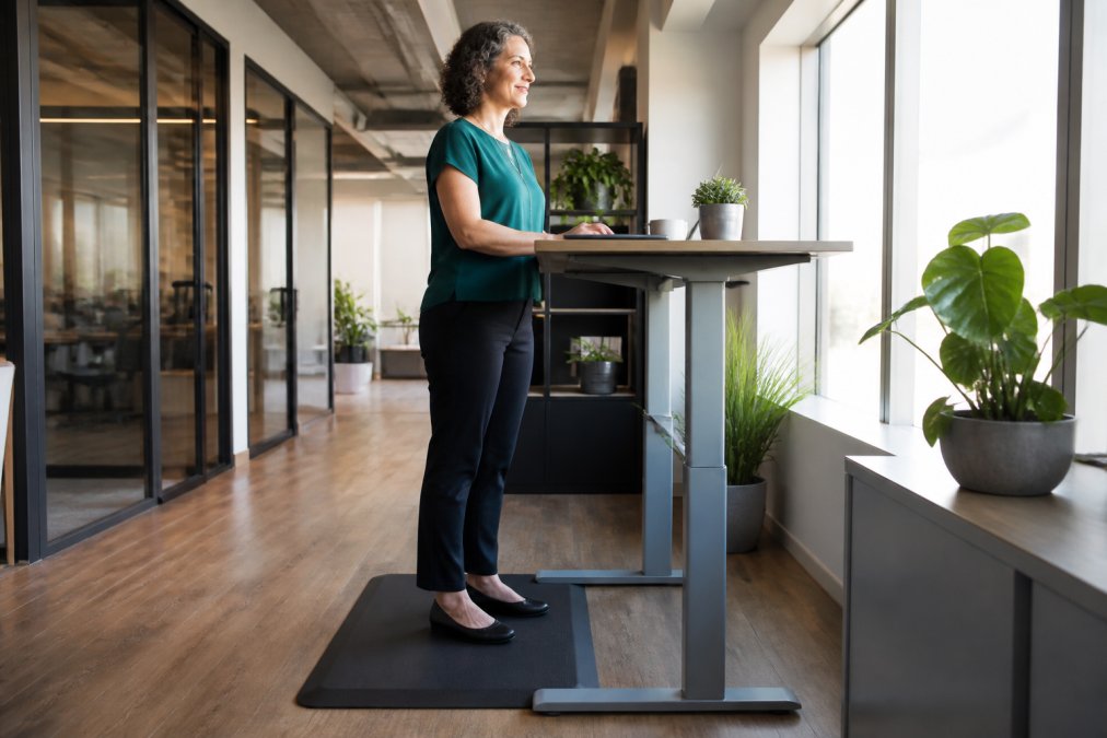 A woman standing on an anti-fatigue mat at a standing desk in a modern office, wearing supportive shoes, with good posture and a relaxed expression