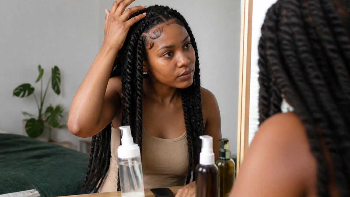 A woman with goddess braids applying lightweight mousse to her braid roots using her fingertips, sitting at a vanity with a mirror and product bottles visible, soft natural lighting