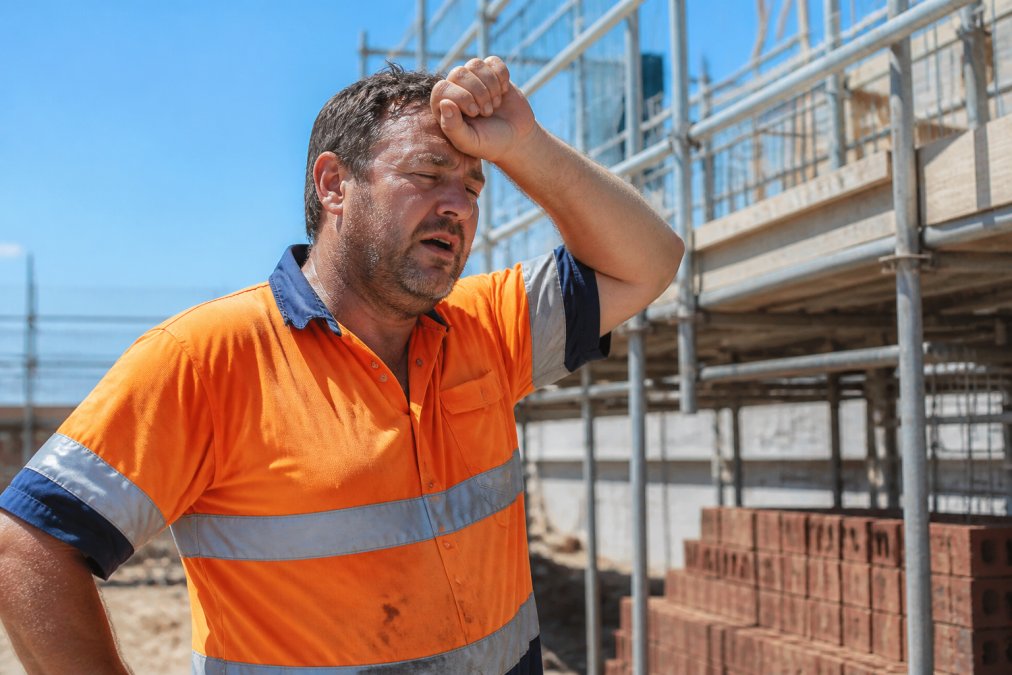 Construction worker on a hot job site wiping sweat from forehead, appearing fatigued in bright sunlight with visible signs of heat stress, wearing high-visibility gear