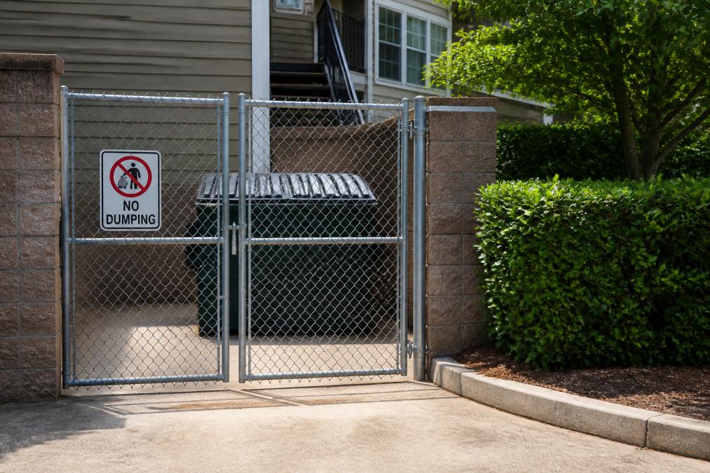 Locked metal dumpster enclosure with a chain-link gate and a no dumping sign behind a rental property