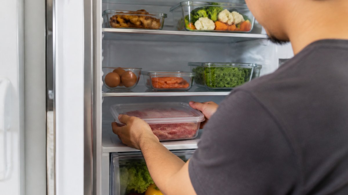 A person placing a sealed container of raw meat on the bottom shelf of a refrigerator, with cooked foods stored on upper shelves, showing clear separation