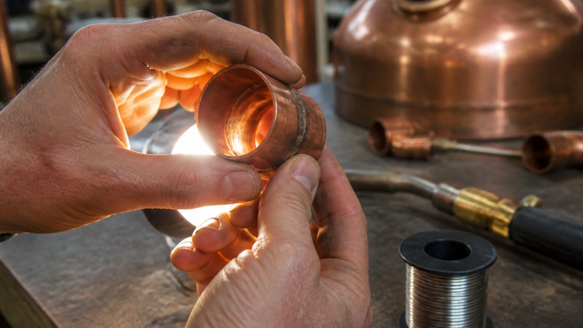 Close-up of a person's hands holding a copper pipe joint up to a light, inspecting the soldered seam for gaps or rough texture, with a soldering torch and lead-free solder wire visible on a workbench in the background