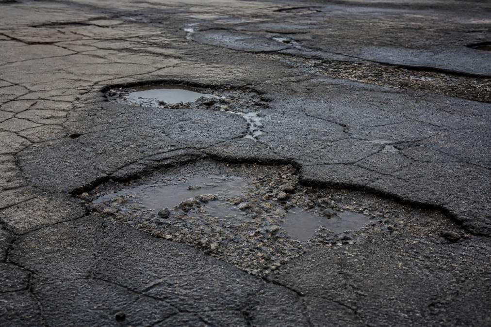 Close-up of deteriorated commercial parking lot surface showing multiple potholes, water pooling in cracks, and spalling asphalt under bright daylight